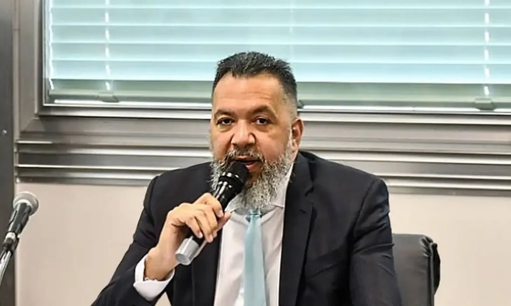 Homem de terno e barba fala ao microfone sentado em plenário