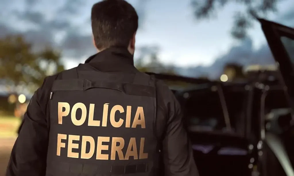 Agente de colete com a inscrição Polícia Federal visto de costas ao lado de viatura