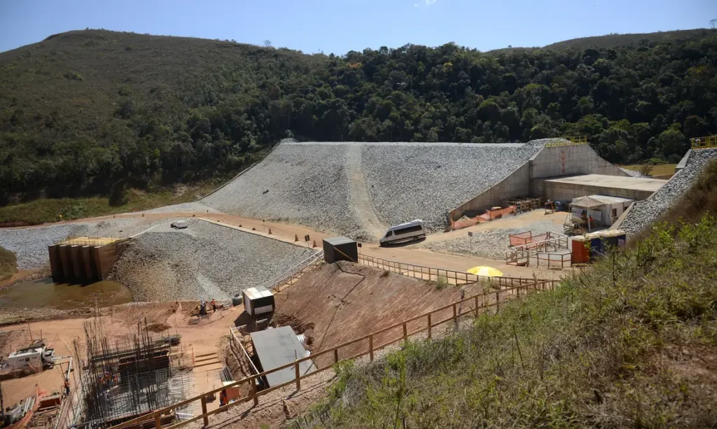 Vista de barragem de rocha em construção entre morros cobertos de mata